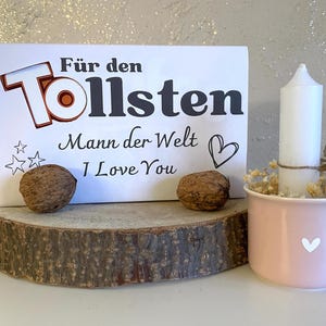 May include: White card with black text that reads "Für den Tollsten Mann der Welt I Love You" with a red heart and two stars. The card is sitting on a wooden slice with two walnuts. A white candle is in a pink mug with a white heart.