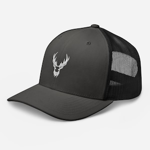Può includere: Un cappello da camionista nero e grigio con un logo di testa di cervo ricamato in bianco e nero. Il cappello ha una parte posteriore in rete nera.