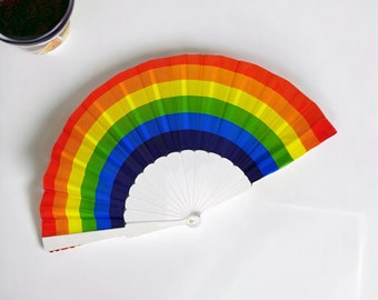May include: A rainbow-colored hand fan with a white handle. The fan is folded and has a rainbow design with red, orange, yellow, green, blue, indigo, and violet stripes.