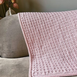 Op de afbeelding: Een zacht, roze gehaakt deken met een textuurpatroon. Het deken is gevouwen en over een bank gelegd.