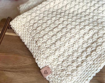 May include: A cream-colored crocheted textile featuring a repeating textured bobble stitch pattern. A small, square, light brown faux leather tag is attached to the corner, embossed with a simple line-art heart icon containing a yarn ball design.