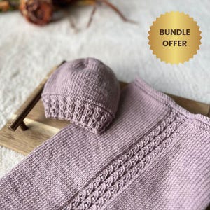 Op de afbeelding: Een gebreide lavendelkleurige baby set, bestaande uit een muts en een deken. De deken heeft een kabelgebreid ontwerp. De set wordt gepresenteerd op een houten dienblad, met een gouden "BUNDLE OFFER" sticker op de achtergrond.