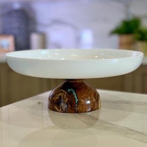 May include: A white serving bowl with a wide, shallow design, elevated on a pedestal base. The base is crafted from polished wood, featuring a natural grain pattern and a turquoise accent. The bowl is ideal for displaying fruit or decorative items.