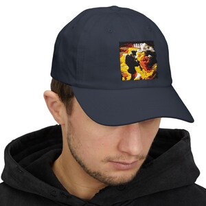 May include: A navy blue baseball cap with a square patch featuring a yellow and orange graphic of a person in a dark suit. The patch has the word "SALOON" in white text.