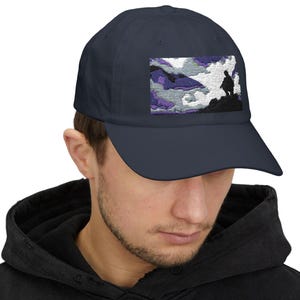 May include: Navy blue baseball cap with a rectangular patch featuring a silhouette of a person on a hill, set against a cloudy sky with purple and white hues. The cap is made of a soft fabric.