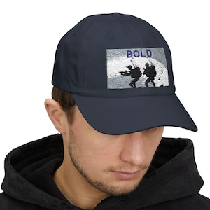 May include: A navy blue baseball cap with a white and gray graphic of soldiers on a mountain with the word "BOLD" in blue text.