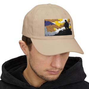 May include: A beige baseball cap with an embroidered image of a person standing on a mountain with a sunset in the background.