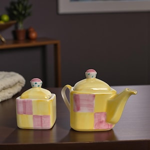 May include: A yellow ceramic teapot and sugar bowl set. The teapot and sugar bowl are decorated with pink and yellow checkered patterns. The lids are topped with pink and green accents. The set is on a dark brown surface.
