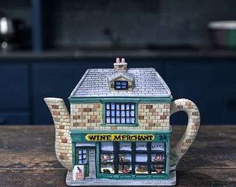 May include: A novelty teapot shaped like a brick building with a green and yellow sign that reads "Wine Merchant". The teapot has a handle and spout, and the building has a detailed facade with windows and a chimney.