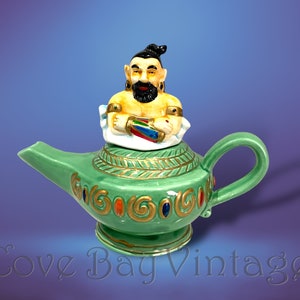 Puede incluir: Una lámpara de genio de cerámica verde con asa y boquilla, con una figura de genio en la parte superior. La lámpara está decorada con detalles dorados y joyas de colores. El genio tiene barba y turbante negros.