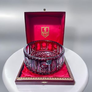 Può includere: Una ciotola di cristallo rosso rubino con intricati motivi intagliati, presentata in una scatola foderata in velluto bordeaux. La ciotola presenta un motivo a stella sulla base e un motivo ad arco ripetuto sui lati. La scatola ha un bordo dorato.