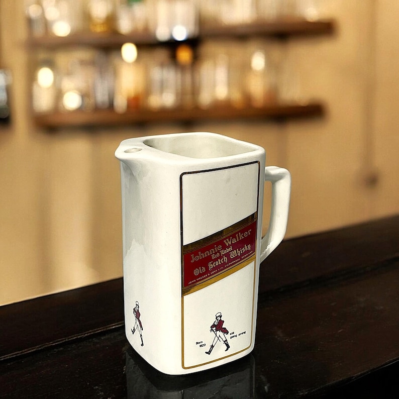 Peut inclure: Pichet en c&eacute;ramique blanche avec une &eacute;tiquette Johnnie Walker Red Label Old Scotch Whisky. L'&eacute;tiquette pr&eacute;sente un design rouge et or avec un homme marchant en manteau rouge.