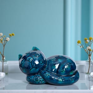 May include: A teal ceramic cat figurine in a sleeping position, decorated with a floral pattern. The figurine is placed on a white surface, with a light blue backdrop. The cat's design features a glossy finish.