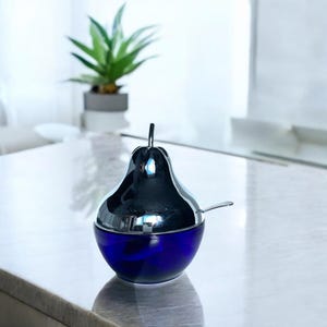 May include: A pear-shaped sugar bowl with a shiny silver lid and a deep blue glass base. A small silver spoon rests on the rim. The bowl is on a white surface, with a plant in the background.