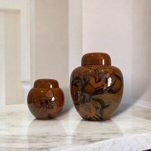May include: Two glossy brown ceramic jars with lids, featuring a hand-painted floral motif in darker brown and black tones. The jars are rounded with a smooth finish, showcasing a consistent colour palette and decorative botanical patterns.