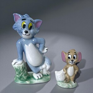 May include: Two ceramic figurines of Tom and Jerry. Tom, a blue cat with yellow eyes, is posed on a green base. Jerry, a brown mouse with pink ears, holds a bag with "TNT" on it. Both are smiling.