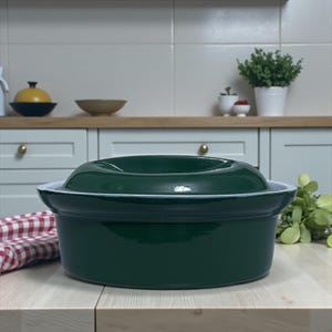 Może przedstawiać: Ciemnozielona ceramiczna forma do zapiekania z pokrywką, ustawiona na jasnej drewnianej powierzchni. Naczynie ma białą obwódkę i znajduje się w otoczeniu kuchennym. Po lewej stronie widoczna jest czerwono-biała kraciasta ściereczka.