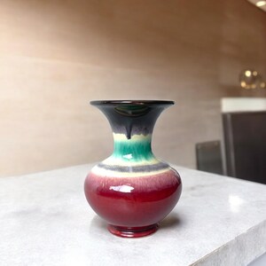 May include: A small ceramic vase with a flared rim and a bulbous body. The glaze features a gradient of colors, transitioning from a deep black at the top, through bands of bright green and white, to a rich, glossy red at the base of the vessel.