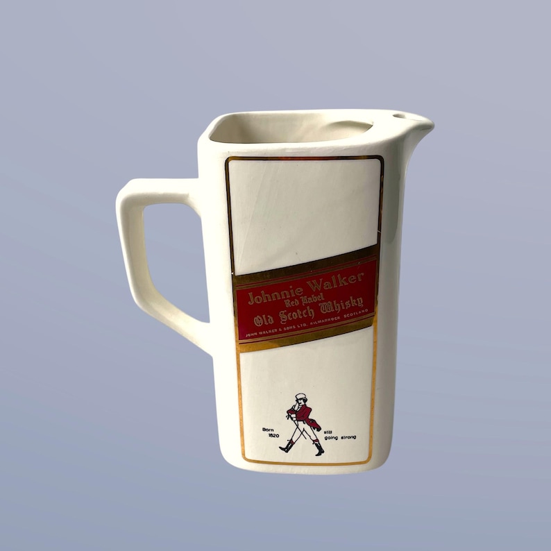 Peut inclure: Pichet en c&eacute;ramique blanche avec une bordure dor&eacute;e et une &eacute;tiquette Johnnie Walker Red Label Old Scotch Whisky. L'&eacute;tiquette est rouge avec des lettres dor&eacute;es et repr&eacute;sente un homme en manteau rouge marchant avec un b&acirc;ton. Le texte sur l'&eacute;tiquette indique "Keep Walking" et "Still Going Strong".