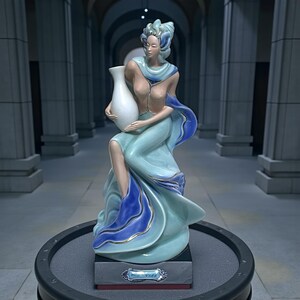 Puede incluir: Estatua de cerámica de una mujer con un vestido fluido turquesa y azul, sosteniendo un jarrón blanco. La estatua tiene una expresión serena y está adornada con detalles dorados. La base de la estatua tiene la inscripción "Se Vida".