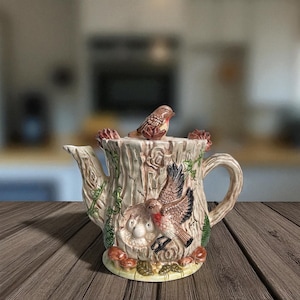 May include: A ceramic teapot with a tree trunk design, featuring a bird perched on top and two birds in flight. The teapot has a cream-coloured base with brown, green, and red accents. The teapot is approximately 20 cm tall.