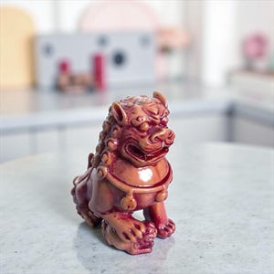 May include: A small, glossy, reddish-brown figurine of a guardian lion, also known as a foo dog. The sculpture features intricate carved details on the mane, a wide open mouth, and a collar with small bell-like ornaments around its neck.