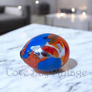 Puede incluir: Un pisapapeles de vidrio abstracto y colorido. El objeto en forma de huevo presenta una vibrante mezcla de tonos azules, naranjas, rojos y blancos. El logotipo de Cove Bay Vintage es visible en la esquina inferior derecha.