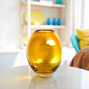 May include: A golden-yellow, oval-shaped glass vase. The vase has a rounded body and a slightly flared opening. It sits on a white surface, reflecting the light. The background includes a wooden shelf with decorative items and a glimpse of a living room.