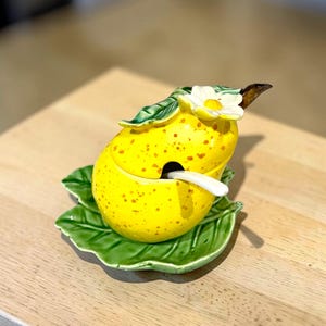 May include: A yellow ceramic jar shaped like a lemon with brown speckles, featuring a lid topped with a white flower and green leaves. The jar sits on a matching green leaf-shaped saucer and includes a small white spoon tucked into the side.