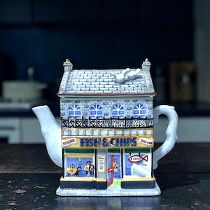 Peut inclure: Théière en céramique en forme de magasin de fish and chips. Le bâtiment a un toit en briques grises, des fenêtres cintrées et l'inscription "FISH & CHIPS" au-dessus de la devanture. La théière est blanche avec une anse et un bec courbés.