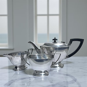 May include: A polished silver tea set comprising a teapot with a black handle and lid, a creamer, and a sugar bowl. The set is arranged on a marble surface, with a window visible in the background.