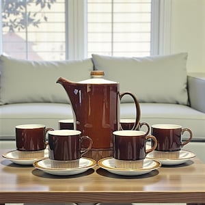 Puede incluir: Un juego de café de cerámica marrón con cafetera, tazas y platillos a juego. Las tazas y la cafetera tienen el interior blanco. El juego está dispuesto sobre una mesa de madera.