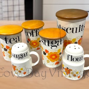 Könnte beinhalten: Satz von sechs Vintage-Küchenbehältern mit Blumendesigns und Holzdeckeln. Die Behälter sind mit "Tee", "Kaffee", "Zucker", "Kekse", "Mehl" und "Zucker" beschriftet.