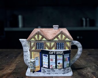 May include: A ceramic teapot shaped like a quaint gift shop. The teapot features a brick chimney, a brown roof, and a storefront with the words "Gift Shop." The handle and spout are designed to resemble brickwork. The shop has an open door and a sign that says "Open".