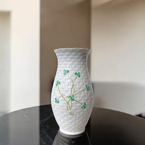 Puede incluir: Un jarrón de cerámica blanca con una textura tejida y un borde festoneado. El jarrón presenta un diseño de trébol verde. El jarrón está sobre una superficie negra.
