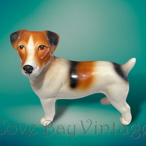Puede incluir: Figura de cerámica de un perro Jack Russell Terrier. El perro tiene un cuerpo blanco con manchas de pelaje negro y marrón. El perro está de pie sobre sus cuatro patas con la cola levantada. La figura tiene un acabado brillante.