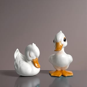 Puede incluir: Dos figuras de pato de cerámica blanca. Un pato está en posición de descanso con los ojos cerrados y un pico naranja. El otro pato está erguido con un pico y patas naranjas y ojos negros. Los patos están sobre una superficie reflectante.