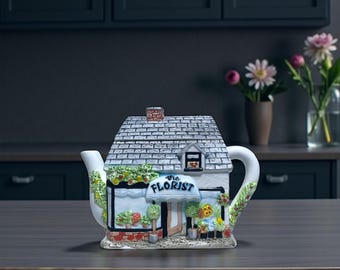 May include: A ceramic teapot shaped like a quaint florist shop. The white building has a gray shingled roof, a small window, and a sign that reads "The Florist." Colorful flowers and greenery decorate the shop's facade and handle.
