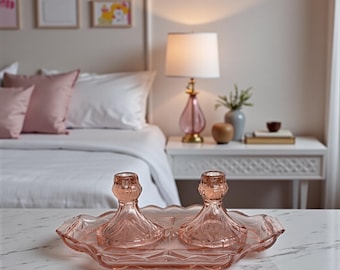 May include: A pair of pink glass candle holders on a matching tray, set on a marble surface. The candle holders have a detailed, textured design. The tray has a scalloped edge and geometric pattern. The background includes a bed and a bedside table with a lamp.