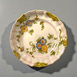 Può includere: Un piatto in ceramica bianca con bordo smerlato, decorato con un motivo floreale. I fiori sono gialli e blu, con foglie e steli verdi. Il piatto ha un bordo rosa e una finitura lucida.