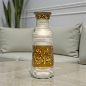 Könnte beinhalten: Eine hohe Keramikvase mit ausgestelltem Rand und zylindrischem Körper. Die Oberfläche zeigt horizontale weiße und cremefarbene Streifen sowie ein markantes Mittelband aus senfgelber Glasur mit einem erhabenen, geschwungenen Tropfenmuster.