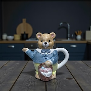 Puede incluir: Una tetera de cerámica caprichosa con forma de oso de peluche, con una chaqueta azul y pantalones amarillos. El oso sostiene una cesta con objetos rosas y blancos. La tetera tiene un asa y un pico blancos, sobre una mesa de madera.