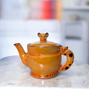 Puede incluir: Una tetera de cerámica naranja con un asa de tapa con forma de oso y un asa con forma de oso. La tetera tiene un acabado brillante y detalles en negro. El asa presenta lunares negros. Un artículo de cocina decorativo.