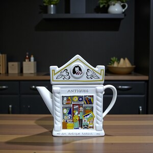 May include: A white ceramic teapot shaped like a building with a removable lid. The front features a colourful illustration of an antique shop interior with the text "ANTIQUES" above and "an antique teapot" printed along the bottom edge.