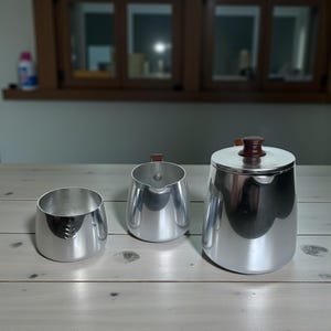 Puede incluir: Un juego de tres piezas de servicio de aluminio cepillado. El juego incluye un cuenco pequeño con un diseño de hoja, una jarra de crema con un asa marrón y un azucarero con un pomo marrón. Las piezas están dispuestas sobre una superficie de madera clara.