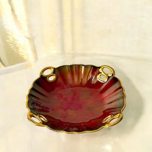 Könnte beinhalten: Eine dekorative Schale mit gewelltem Rand, tiefrotem Innenraum und goldfarbener Verzierung. Das muschelförmige Design hat goldene Griffe. Der Innenraum hat eine glänzende Oberfläche, und die Schale scheint aus Metall zu sein.