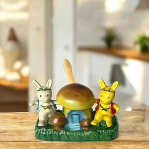 May include: A ceramic mushroom-shaped salt and pepper shaker set. The set features a brown and yellow mushroom house with a blue door, flanked by two bunny figurines. The base is green with grass detailing, and a wooden spoon is included.