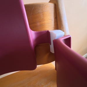 Pode incluir: Um acessório para cadeira de refeição com encosto e barra frontal em plástico liso rosa-escuro, ligados por um mecanismo de bloqueio em plástico branco, montado numa estrutura de cadeira de madeira clara com veios visíveis.