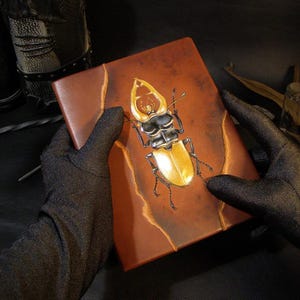 May include: A brown leather journal with a gold beetle design on the cover. The journal is being held by two gloved hands.