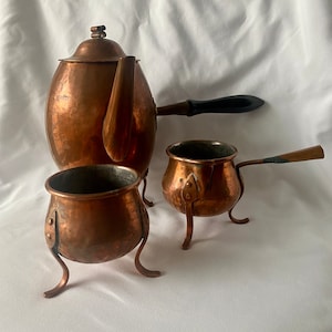 Könnte beinhalten: Ein Set aus drei Kupfertöpfen mit Griffen. Der größte Topf hat einen Ausguss und einen schwarzen Griff. Die beiden kleineren Töpfe haben jeweils drei Beine.